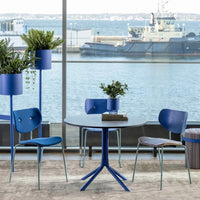 Blue modern chairs and table with circular patterns, complemented by green plants in blue pots, against a water view.