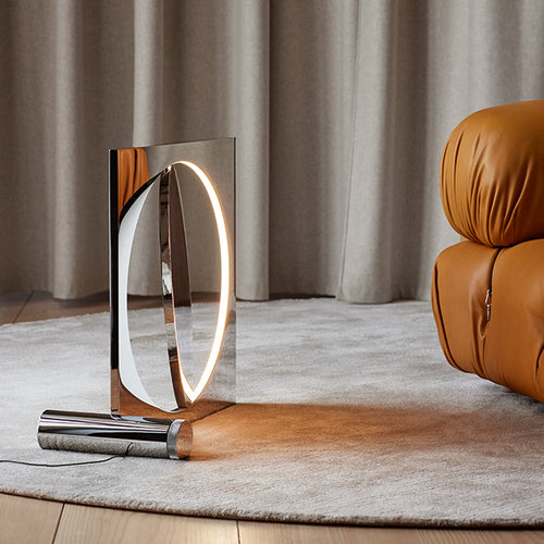 Sleek chrome lamp with a circular light, set against a soft beige rug and warm brown leather chair. Modern and stylish design.