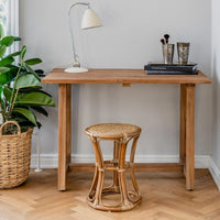 "Wooden desk and rattan stool in natural tones, with a green plant and soft gray wall backdrop."