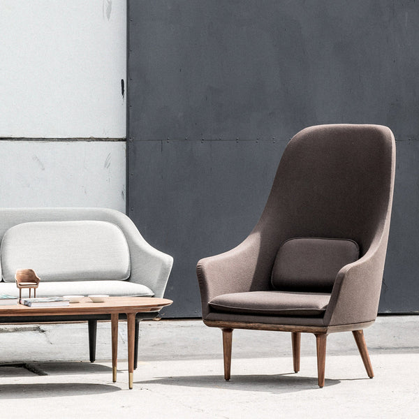 Modern high-back armchair in rich brown fabric, paired with a light gray curved sofa, set against a minimalist backdrop.