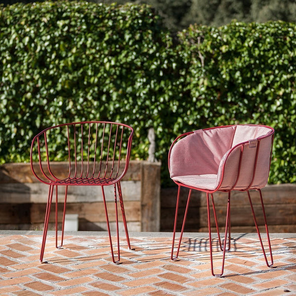 "Red wire chair and blush upholstered chair on a rustic brick patio, surrounded by lush greenery."