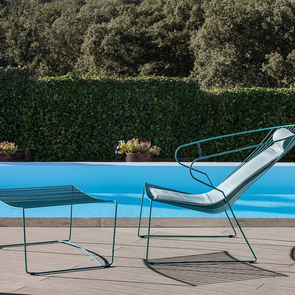 Green metal lounge chair and side table with minimalist design by a pool, featuring a light gray fabric seat.