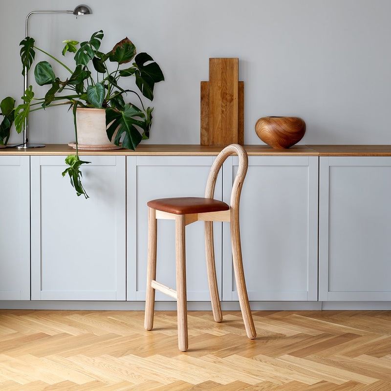 Light wood bar stool with a brown leather seat, set against a soft gray cabinet and warm wooden accents.