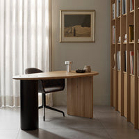 Modern wooden desk with a light oak finish, black cylindrical base, and a patterned chair in muted tones.