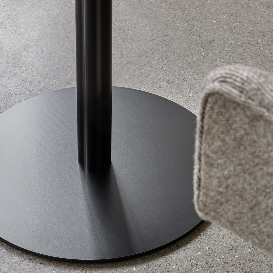 Round black metal base with sleek design, complemented by a textured beige fabric cushion corner. 