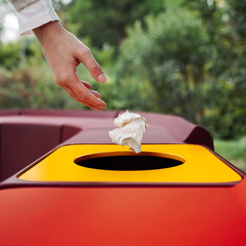 Hand dropping waste into a vibrant red trash can with a bold yellow slot, showcasing a sleek, modern design.