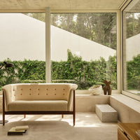 Beige tufted sofa with wooden legs, set against a neutral interior and lush green outdoor backdrop.
