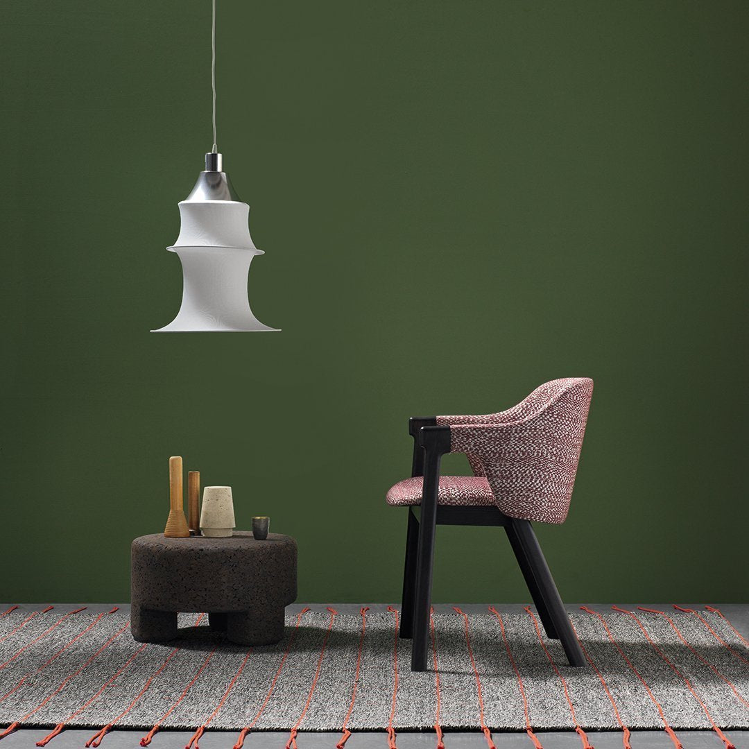 Pink patterned chair, white lamp, black round table, and colorful woven rug against a green background.