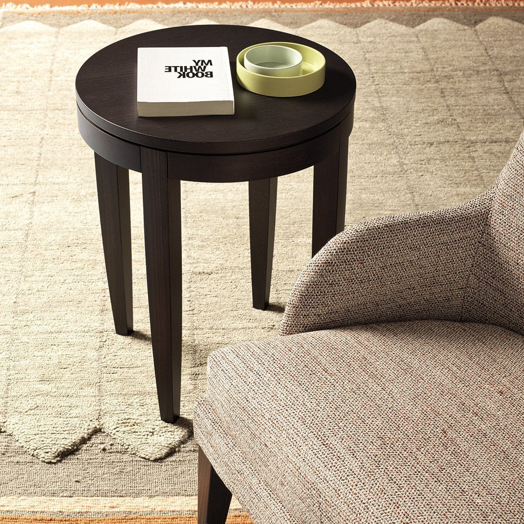 Dark wood round side table with tapered legs, paired with a textured beige and gray armchair. Elegant, minimalist design.