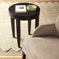 Dark wood round side table with tapered legs, paired with a textured beige and gray armchair. Elegant, minimalist design.
