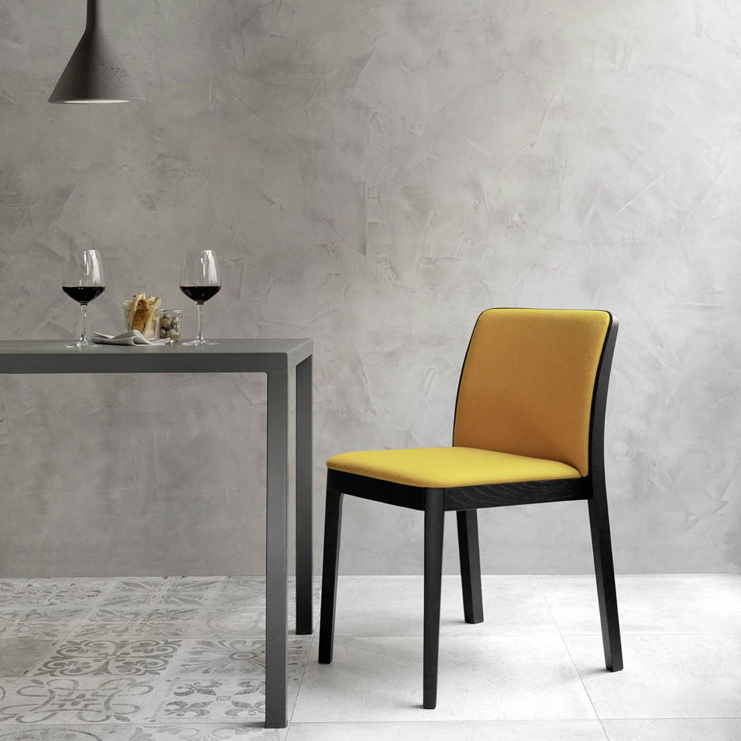 Yellow upholstered dining chair with a sleek black frame, set against a textured gray wall and patterned floor tiles.