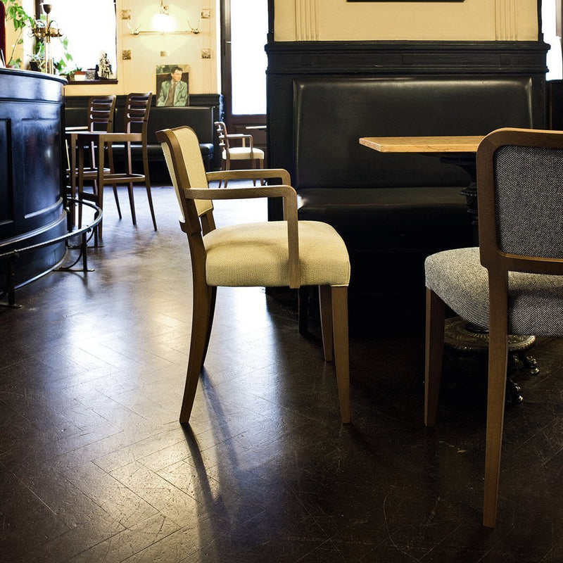 Beige upholstered chair with wooden armrests in a stylish café setting, featuring dark wood floor and varied seating.