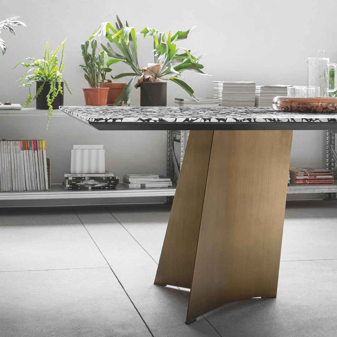 Modern table with a patterned black and white top and a sleek gold base, surrounded by greenery and minimalist decor.