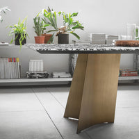 Modern table with a patterned black and white top and a sleek gold base, surrounded by greenery and minimalist decor.