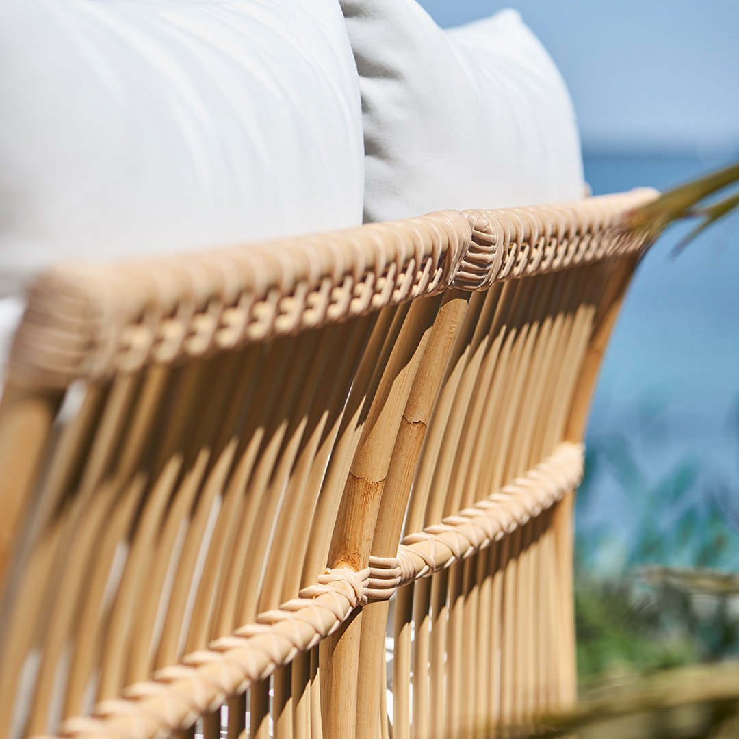 Natural rattan chair back with light beige cushions, showcasing intricate weaving patterns against a serene outdoor backdrop.