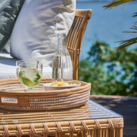 Woven rattan tray with clear drink, vibrant greens, and earthy neutrals, set against a serene blue backdrop.