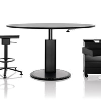 Black round table with sleek surface and cylindrical base, accompanied by black office chair and storage unit.