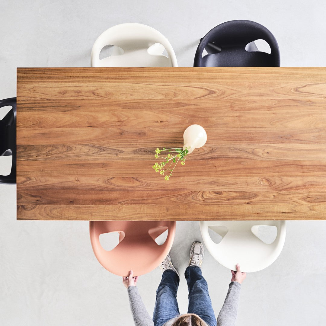 Wooden dining table with earthy tones, surrounded by pastel pink, white, and black chairs against a light gray background.