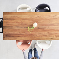 Wooden dining table with earthy tones, surrounded by pastel pink, white, and black chairs against a light gray background.