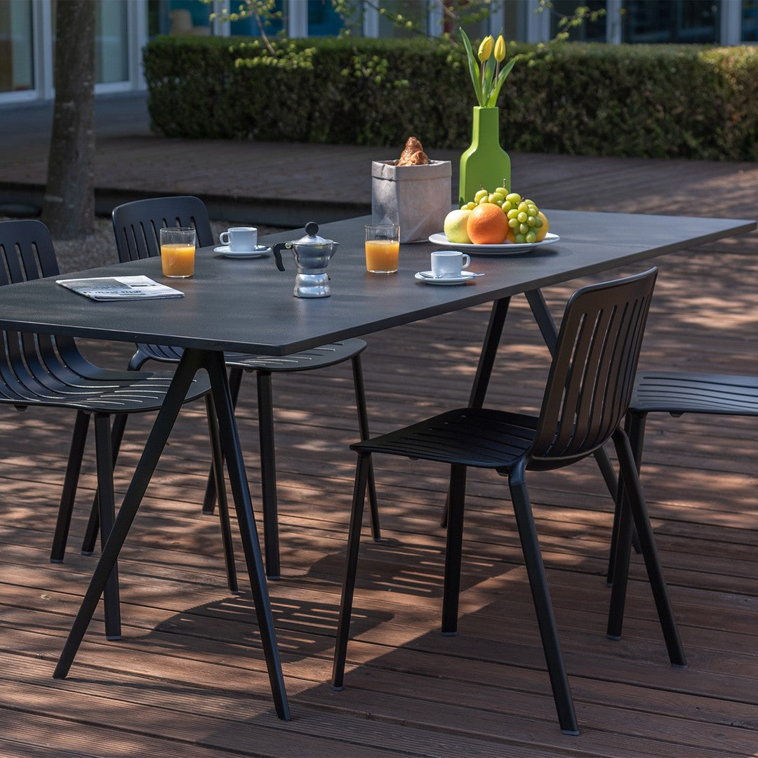 Black outdoor dining table with patterned chairs, vibrant green vase, and colorful fruit accents on a wooden deck.