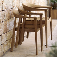 Stacked wooden chairs in a warm brown finish against a textured stone wall, creating a natural, rustic aesthetic.