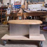 "Modern wooden bench with a smooth light brown top and sleek silver metal base, set in a rustic workshop."