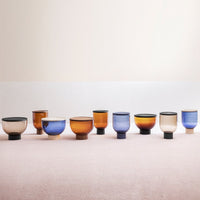 Colorful glass cups in amber, blue, and beige with unique patterns and a modern design displayed on a soft pink background.