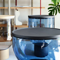 Round coffee tables in clear blue and amber glass with black tops, featuring smooth, modern design and minimal patterns.