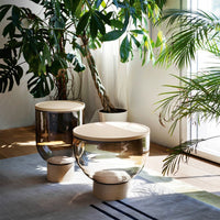Two modern glass side tables with a clear and amber gradient, topped with light wood, surrounded by green plants.