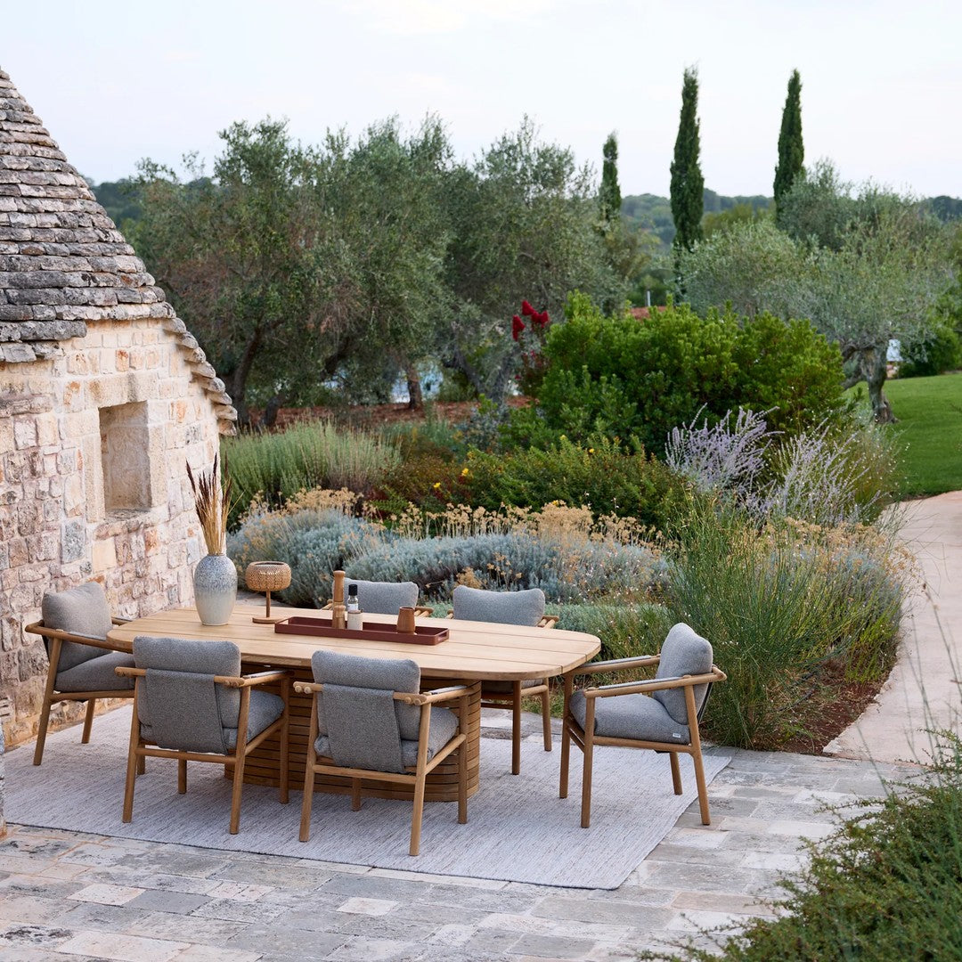 "Natural wood dining table with gray cushions, surrounded by lush greenery and stone pathway in a serene outdoor setting."