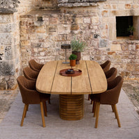 Natural wood oval dining table with warm brown, textured chairs and a beige patterned rug on a rustic stone backdrop.