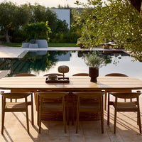 Natural wood dining table with beige chairs, set against a serene poolside green backdrop and soft neutral tones.