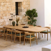 Large wooden dining table with natural finish, paired with light wood chairs and teal cushions against a stone backdrop.