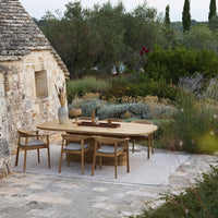 Natural wood dining table with neutral cushions, surrounded by lush green foliage and patterned stone flooring.