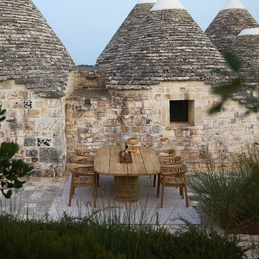 "Natural stone wall with earthy tones, rustic round wood table, and woven rattan chairs amidst greenery."