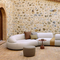 Curved white sofa with brown and mustard cushions, paired with a round brown table, set against a textured stone wall.