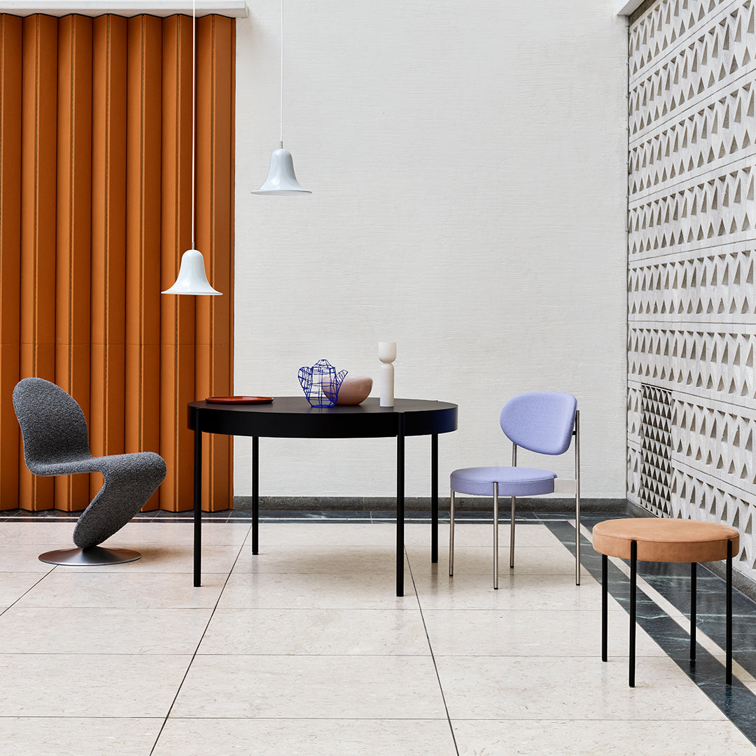 Modern dining room featuring orange curtains, a black round table, lavender chair, gray chair, and patterned wall.