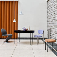 Modern dining room featuring orange curtains, a black round table, lavender chair, gray chair, and patterned wall.