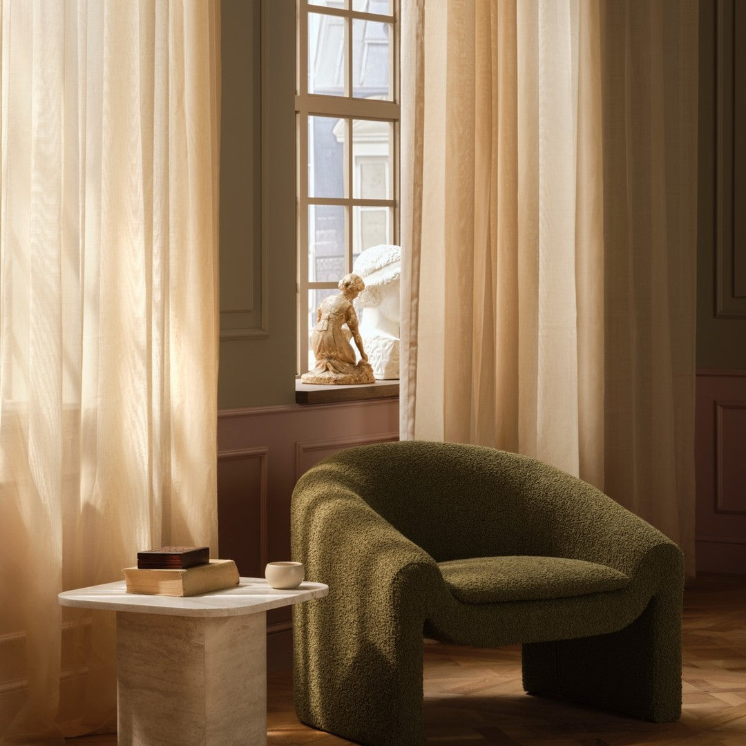 Green curved chair beside a marble table, with soft beige curtains filtering natural light. Elegant and modern design.