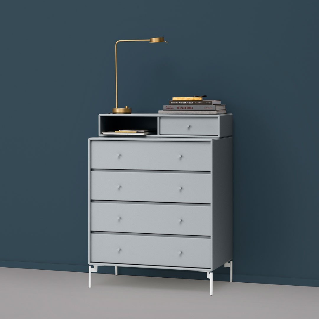 Gray five-drawer dresser with a modern design and sleek white legs against a dark blue wall, topped with a gold lamp.