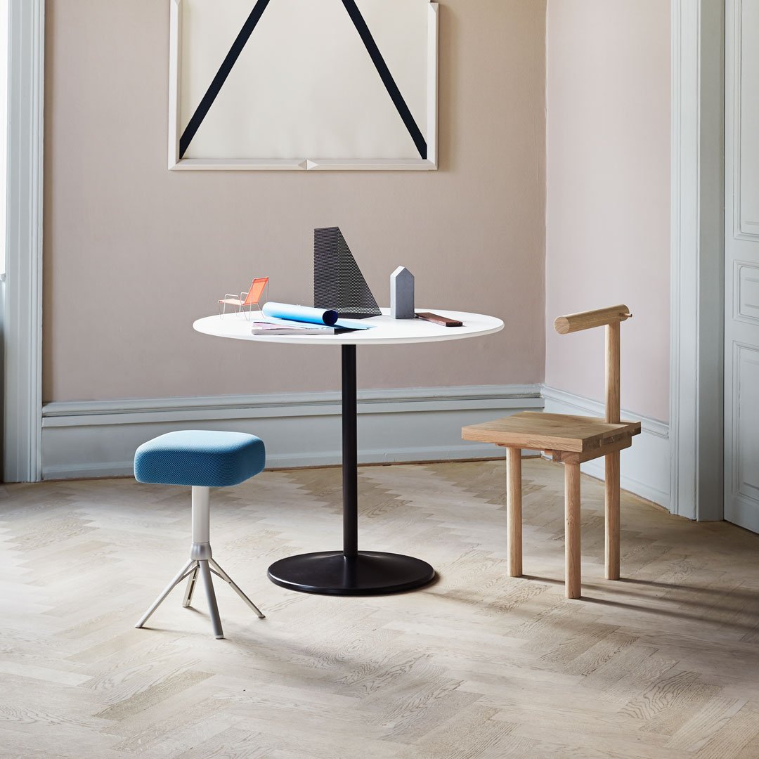 Minimalist round table with a white surface, black base, light wood chair, and blue stool in a soft pastel room.