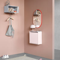Soft pink wall with a light gray shelf and a red-framed mirror. Gray bag and pastel rug add subtle texture to the space.