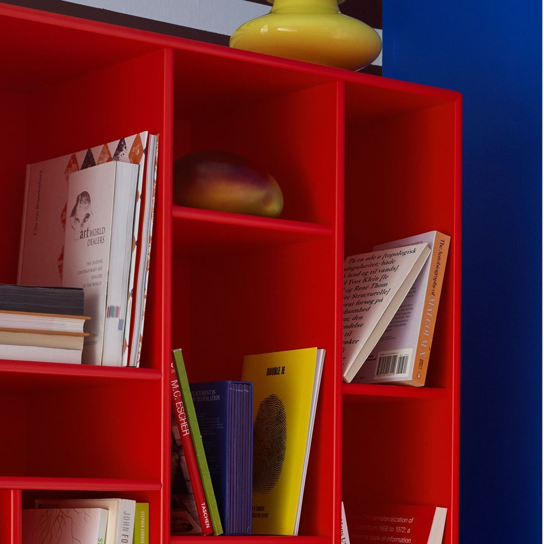Bright red bookshelf with varied colored books and a bold, yellow vase, set against a blue wall.
