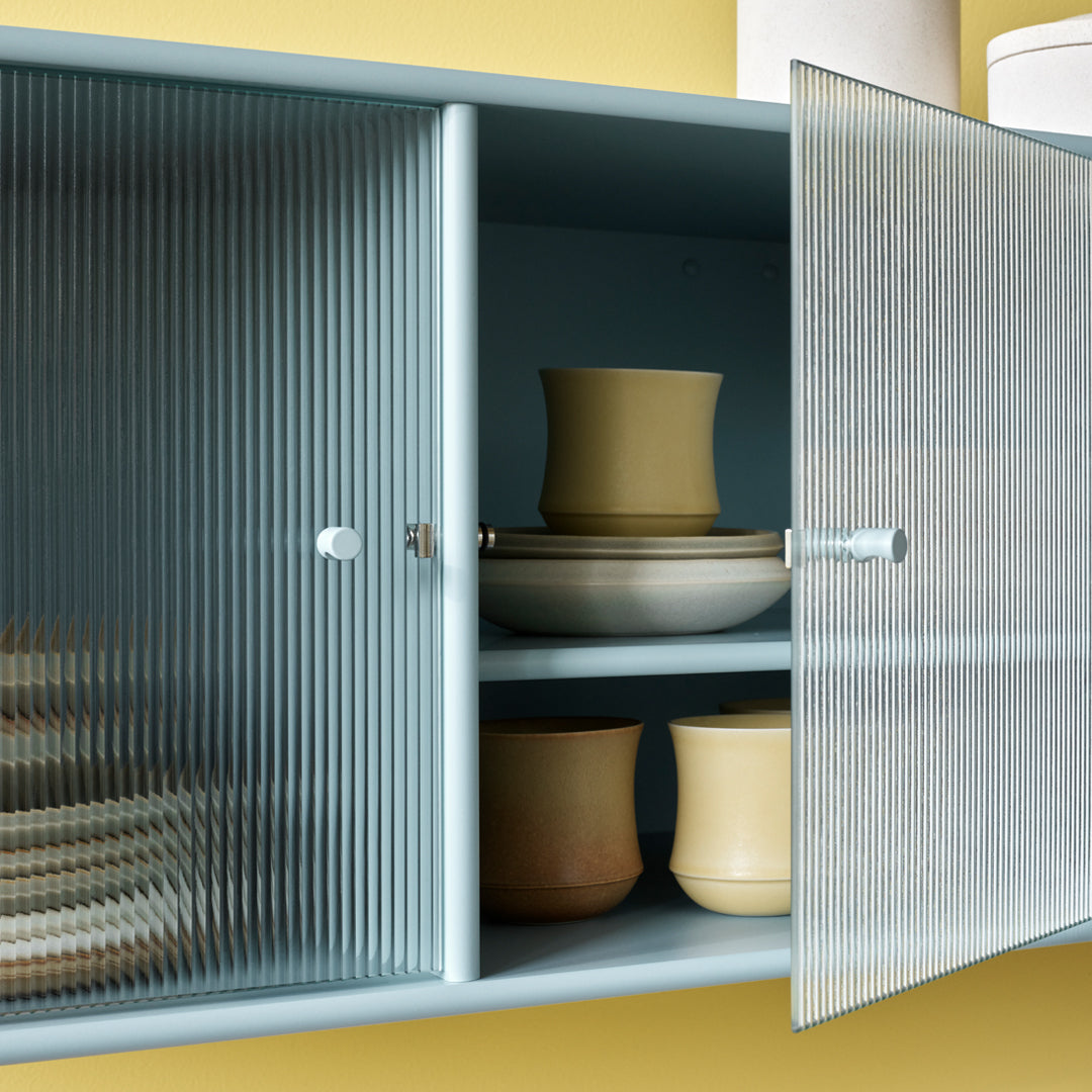 Light blue cabinet with ribbed glass doors, showcasing beige and brown ceramic dishes against a soft yellow backdrop.