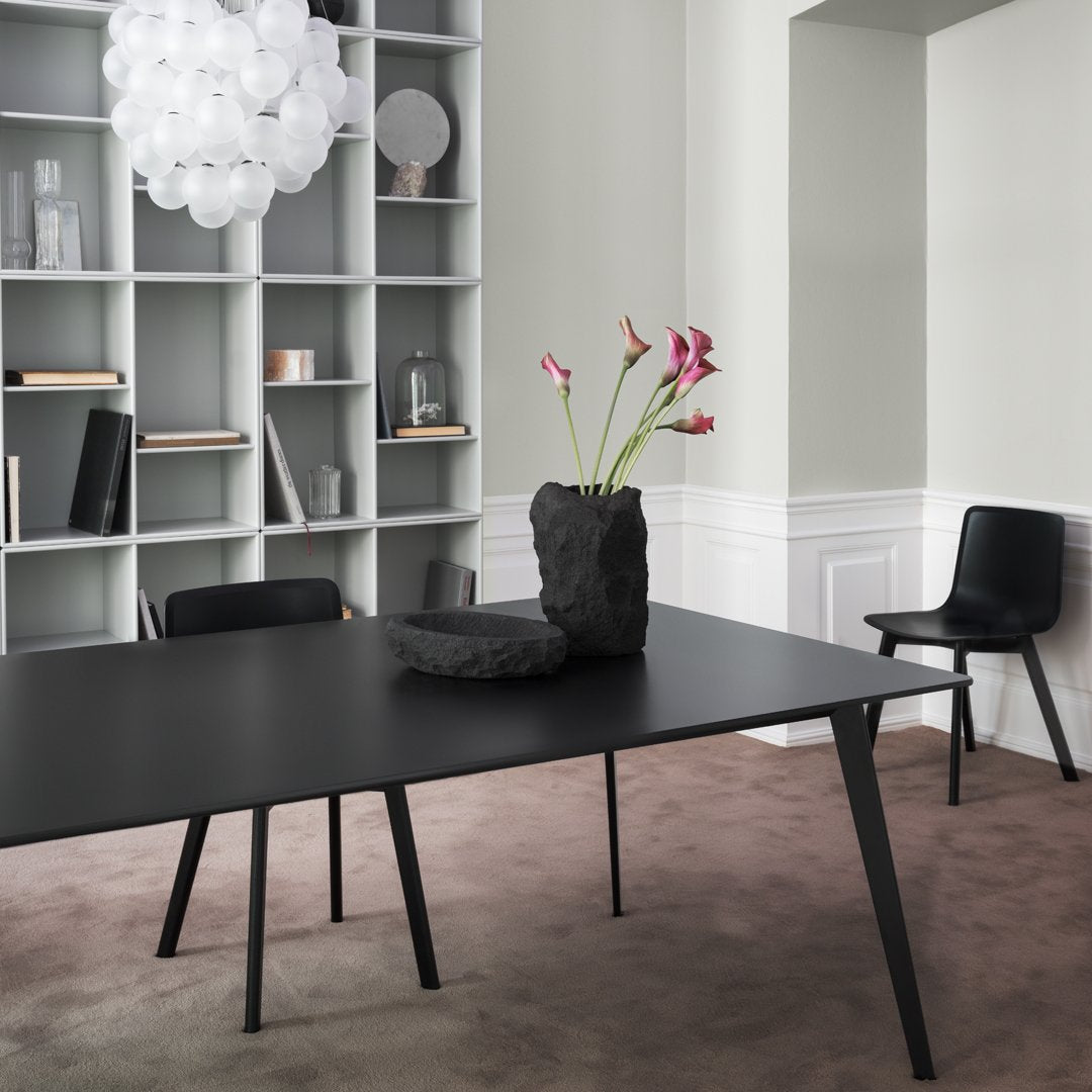 Black modern table, gray shelving, soft pink carpet, and black chair with a textured black vase and pink flowers.