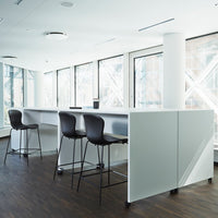 Modern white bar table with black stools in a minimalist setting, featuring large windows and wooden flooring.