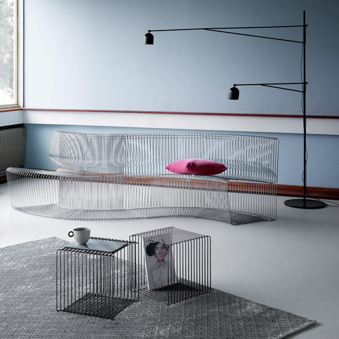 Transparent wire frame sofa with a pink cushion against a minimalist blue wall, featuring black accent tables and lamp.