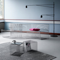 Transparent wire frame sofa with a pink cushion against a minimalist blue wall, featuring black accent tables and lamp.