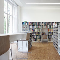 Minimalist white room with beige chairs and brown wooden floor; colorful books arranged on black shelves.