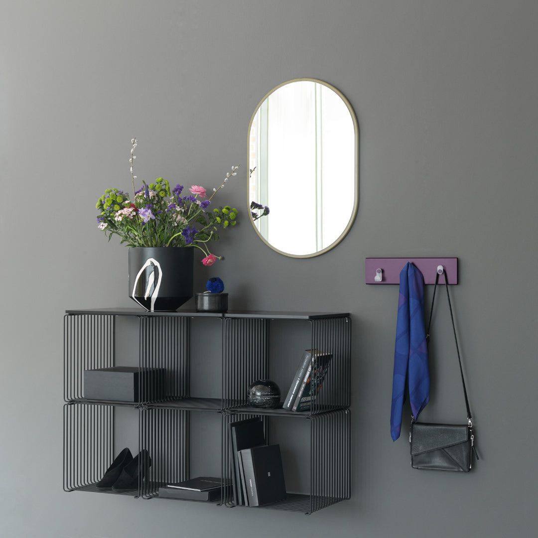 Black wire shelving with a purple hook, green and purple flowers, and a round gold-framed mirror on a gray wall.